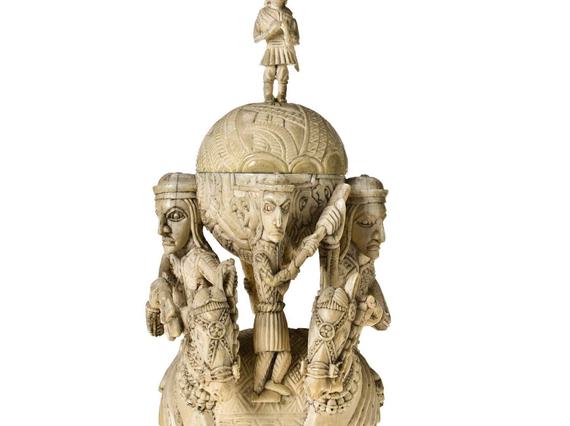 Tall off-yellow ivory sculpture. Two figures on horseback flack two orbs, on atop the other. A small human figure stand between the orbs, another crowns the sculpture.