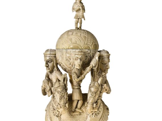 Tall off-yellow ivory sculpture. Two figures on horseback flack two orbs, on atop the other. A small human figure stand between the orbs, another crowns the sculpture.