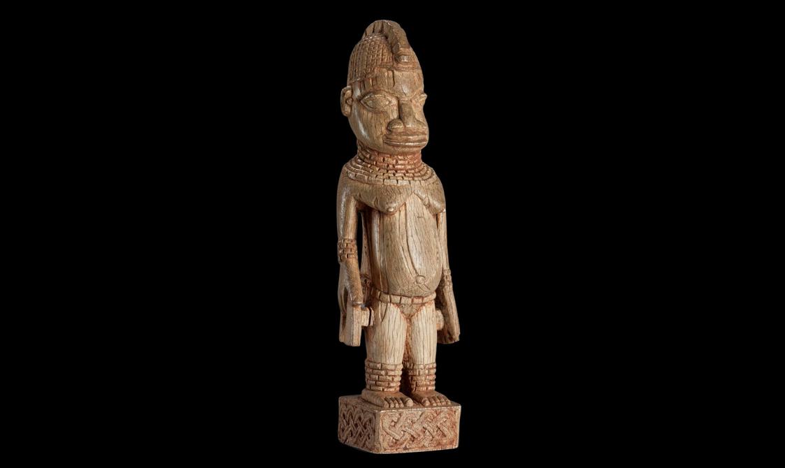 A light brown sculpture of standing a person wearing jewellery around their neck, ankles, and wrists. They stand on a small decorated rectangle.