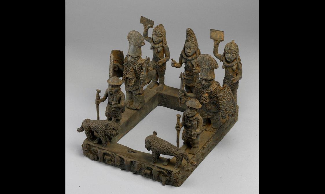 A brass sculpture of various figures and two leopards. The figures wearing elaborate clothes and are placed on top of an open rectangle.