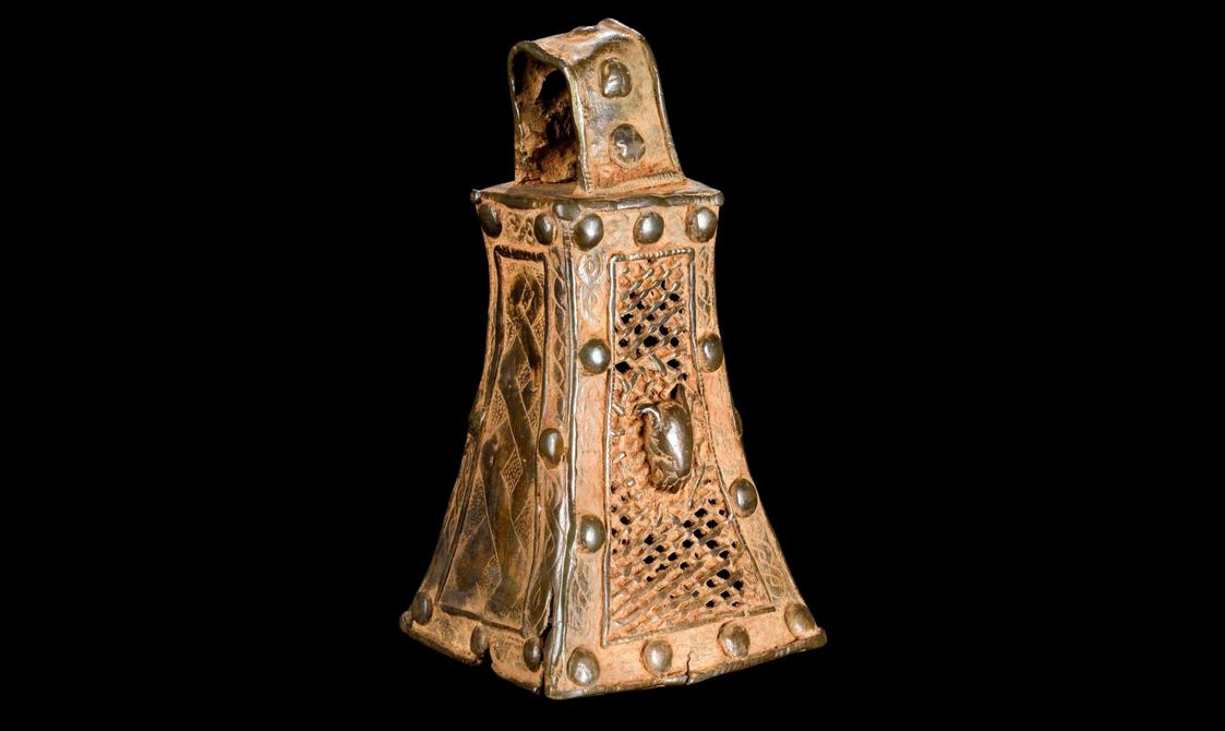 A brass bell with four flat intricately carved sides and a looped handle on top