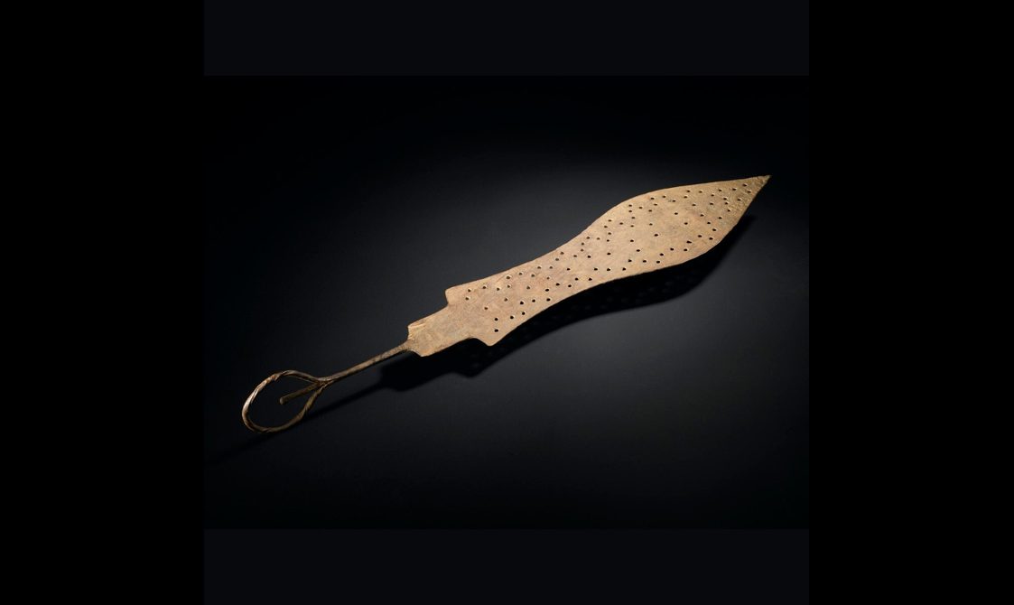 A spear shaped brass object with small holes punched on its surface. It has a thin handle with a loop at the end.