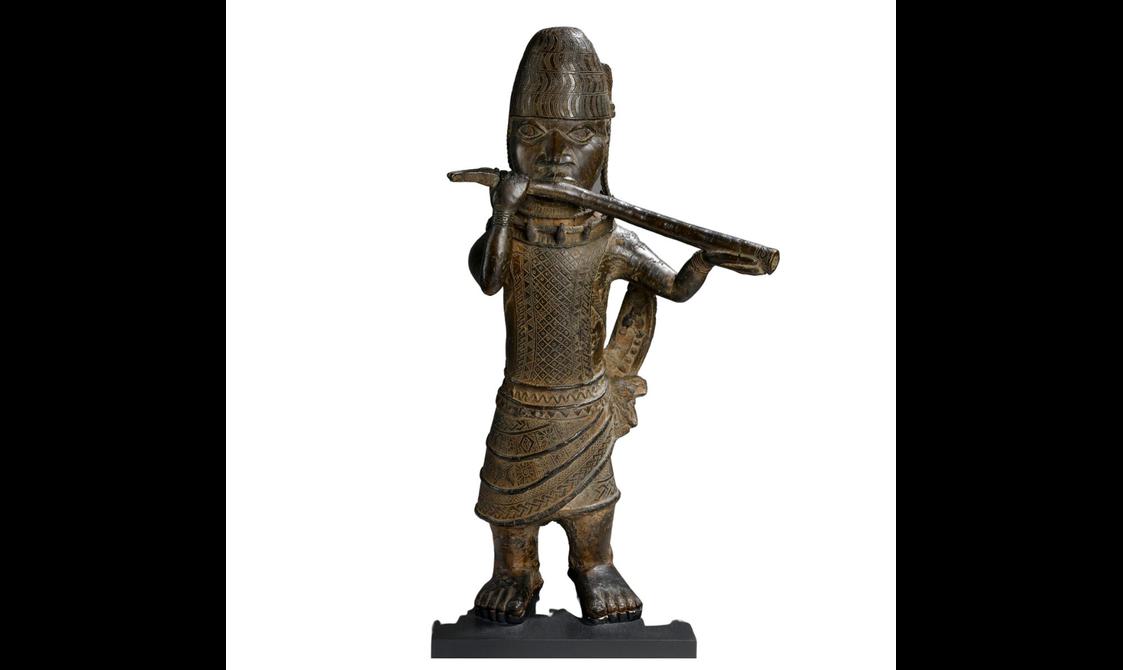 A brown sculpture of a person wearing a wrap skirt with intricate carvings on its body. They are wearing a tall hay and are holding a long horn.