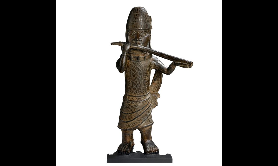 A brown sculpture of a person wearing a wrap skirt with intricate carvings on its body. They are wearing a tall hay and are holding a long horn.