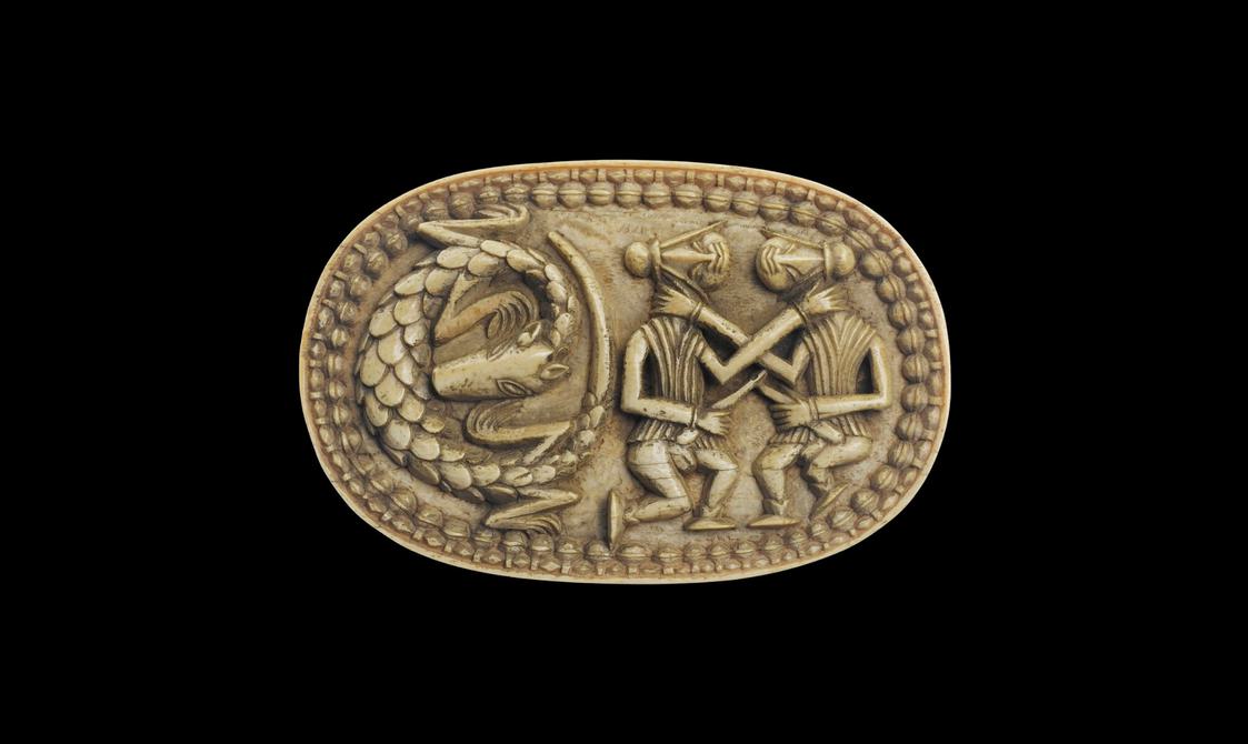 An oval shaped ivory sculpture showing two stylised figures fighting. There is a scaly creature to their left and a boarder of a beaded shape.