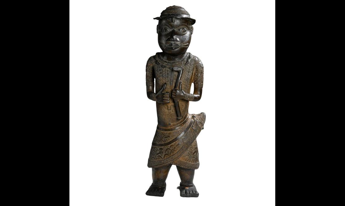 A brown sculpture of a person wearing a wrap skirt with intricate carvings across its body. It is holding a long thin object.