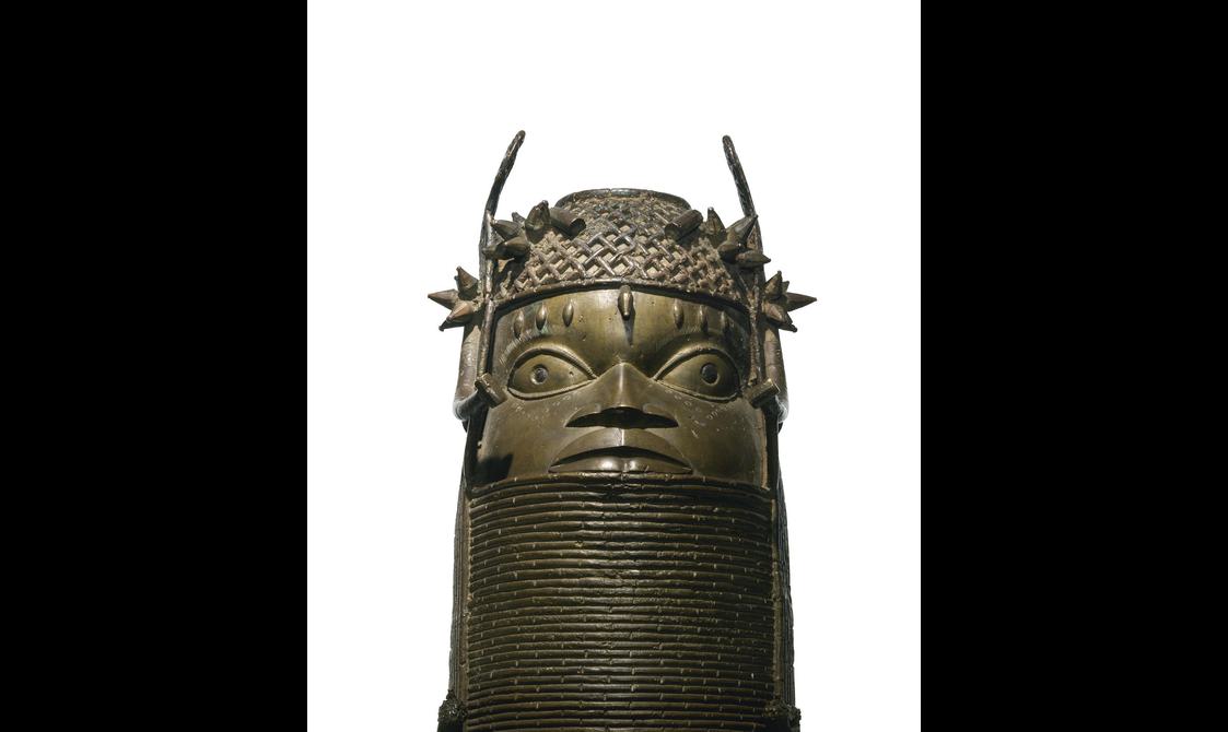 A bronze sculpture of a face with large eyes with a lattice detailed headpiece. The sculpture has a thick base with coiled rings.