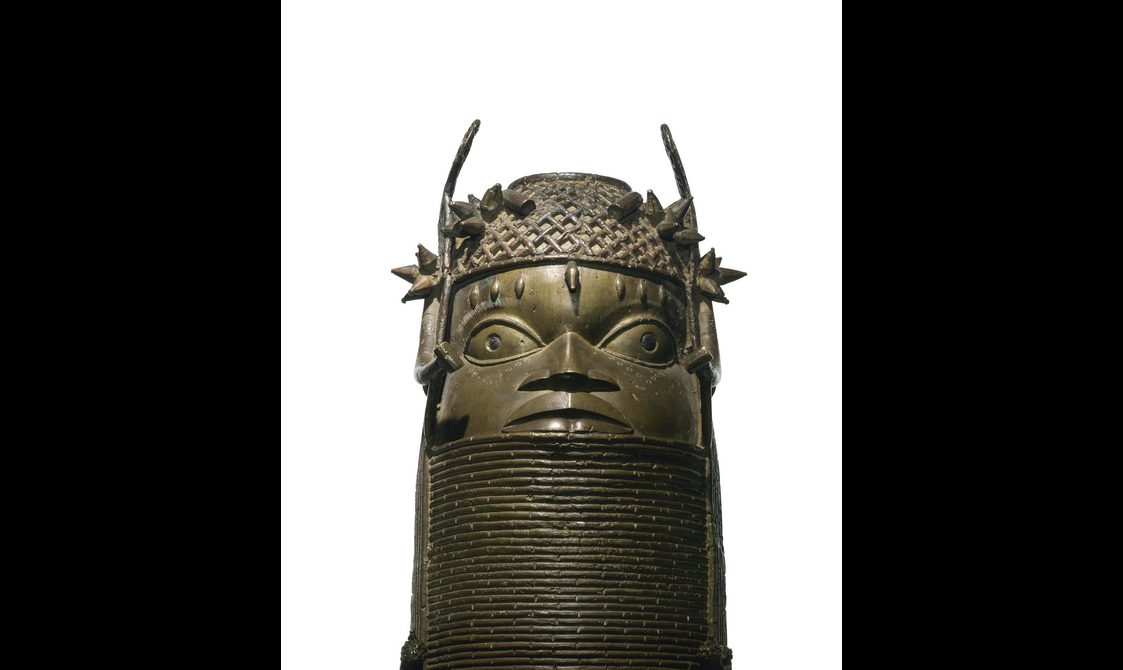 A bronze sculpture of a face with large eyes with a lattice detailed headpiece. The sculpture has a thick base with coiled rings.