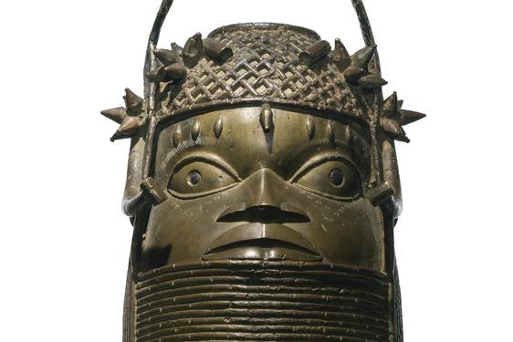 A bronze sculpture of a face with large eyes with a lattice detailed headpiece. The sculpture has a thick base with coiled rings.