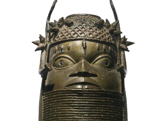 A bronze sculpture of a face with large eyes with a lattice detailed headpiece. The sculpture has a thick base with coiled rings.