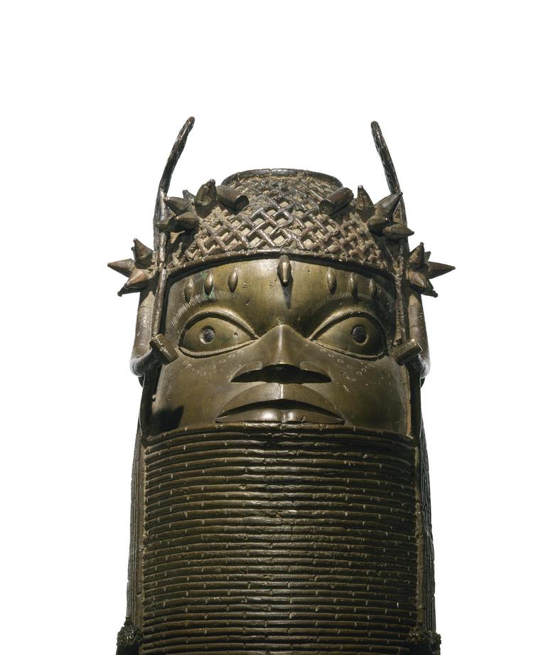 A bronze sculpture of a face with large eyes with a lattice detailed headpiece. The sculpture has a thick base with coiled rings.