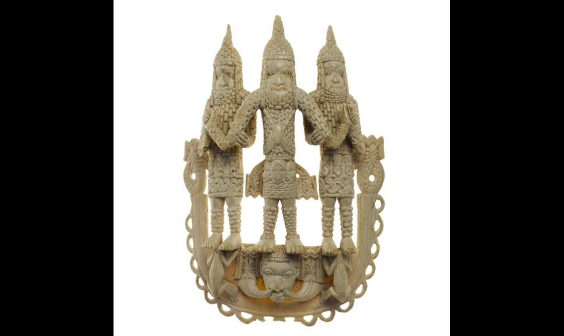 An ivory sculptural object featuring three stylised figures dressed in armour standing on two frogs and a face.