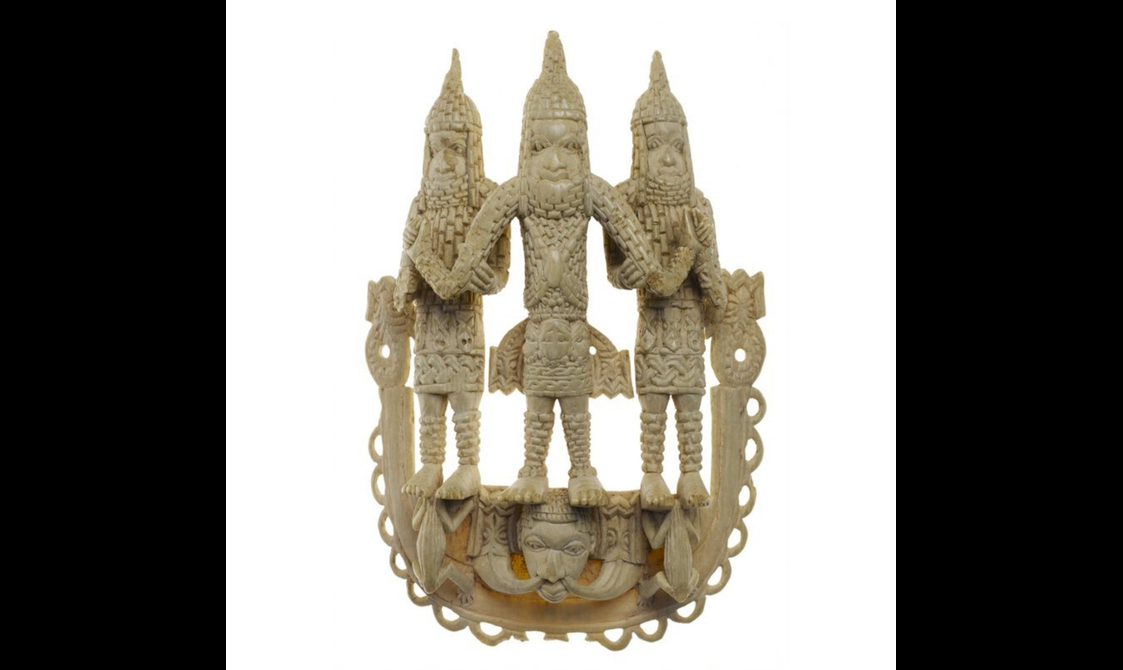 An ivory sculptural object featuring three stylised figures dressed in armour standing on two frogs and a face.