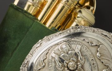 The top of a silver container with a decorative engraving of a saint. In the background are various gold objects held together in a green velvet wrapping.