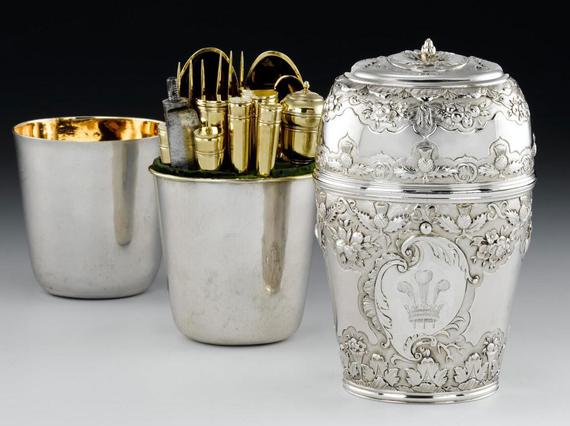 Three silver cylinder shaped boxes, placed behind each other, first box with the lid is decorated in leaf and scottish thistle motifs and a crown with three feathers in the center. Two boxes are plain silver with gilded interior. The middle box contains gold cutlery.
