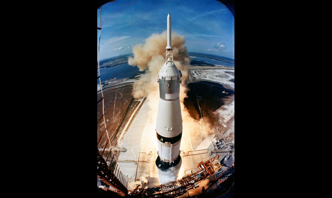 A white rocket launches from its launch pad in a port area, surrounded by smoke.
