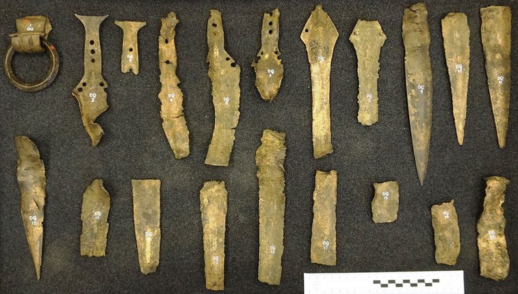 A colour photograph of a collection of twenty brass broken pieces against a grey background. The view is from above. The items include a ring holder and several broken sword pieces.