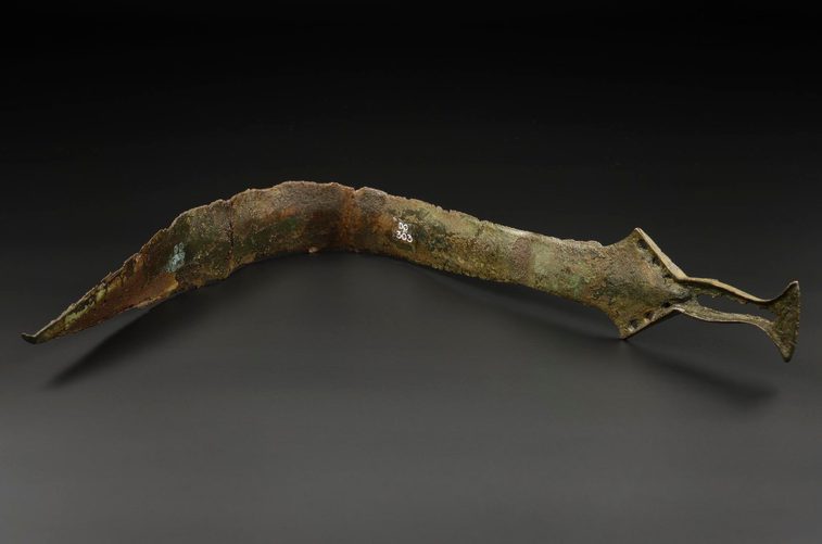 A colour photgraph of a corroded very old bronze bent sword against a grey table background. There is a black 5cm rule indicator underneath the sword to help give scale.