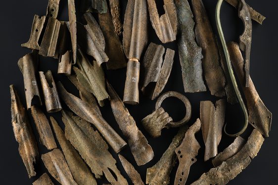 A group of Bronze Age objects including a ring of a large cauldron and fragments of spearheads, swords and dagger blades against a black background.