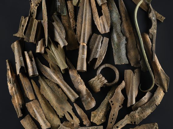 A group of Bronze Age objects including a ring of a large cauldron and fragments of spearheads, swords and dagger blades against a black background.