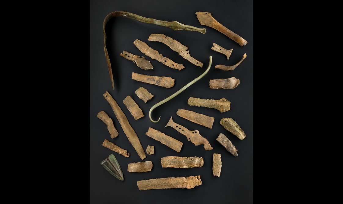 A colour photograph of a pile of very old bronze bone pieces in a pile against a black background. The objects include spearheads, bits of swords, a hook and several sword pieces.