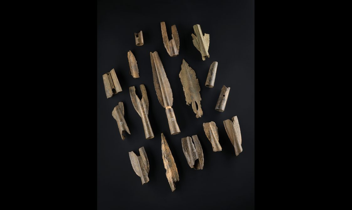 A colour photograph of a pile of very old bronze bone pieces in a pile against a black background. The objects include mostly spearheads.