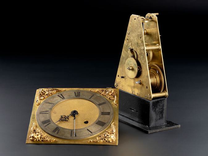 A round clockface on a gold square laying beside a gold triangle with cogs on its surface and its inside. mounted on a rectangle black base.