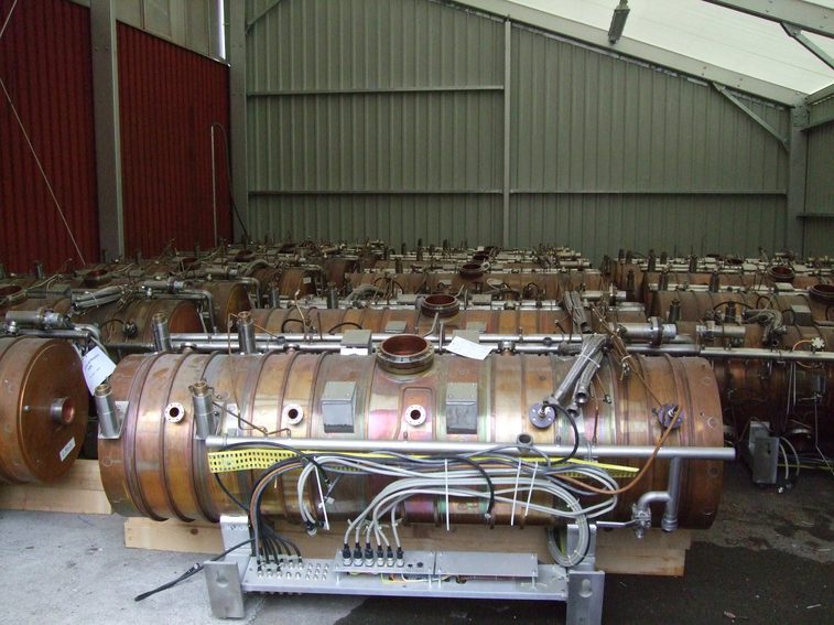 A colour photograph of several copper tubes in a metal industrial shed. Each copper tube sites on a wooden support with a metal frame. They each.have various inlet and outlet holes and many communication wires attached at the bottom.