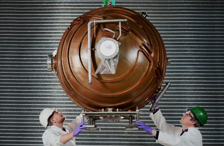 A colour photograph of a large suspended copper sphere. The sphere is ridged and has several metal attachments. At the bottom, either side, are 2 men in safety helmets inspecting the sphere above.