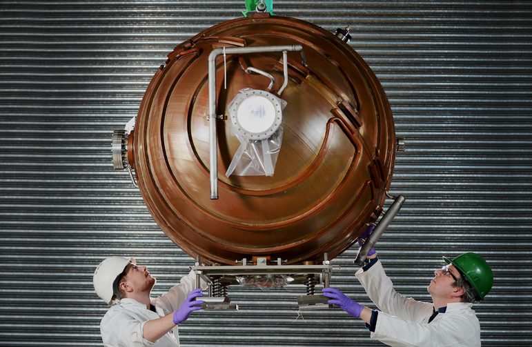 A colour photograph of a large suspended copper sphere. The sphere is ridged and has several metal attachments. At the bottom, either side, are 2 men in safety helmets inspecting the sphere above.