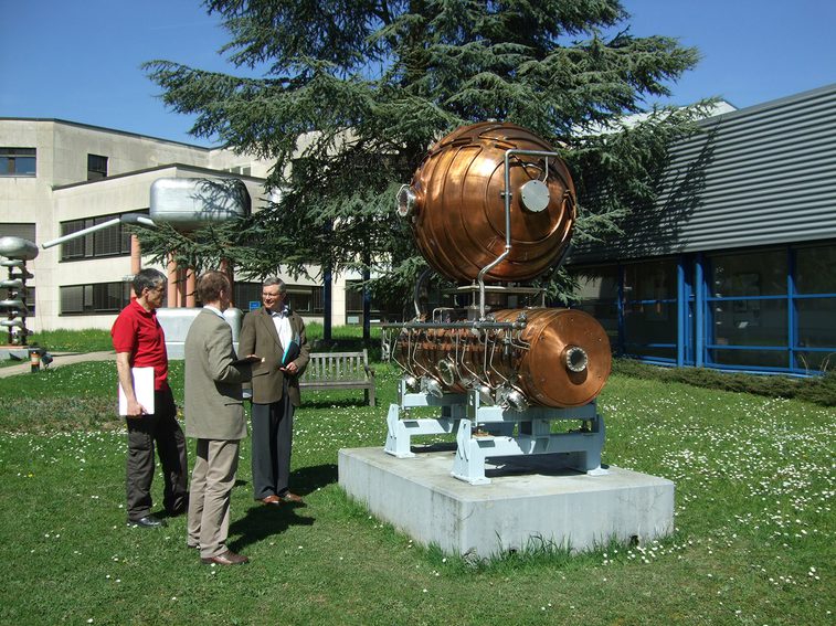 A colour photograph of 3 men in a gardens looking at a copper sculpture mounted on a metal and concrete base. There is a large tree in the background and the side of a building.