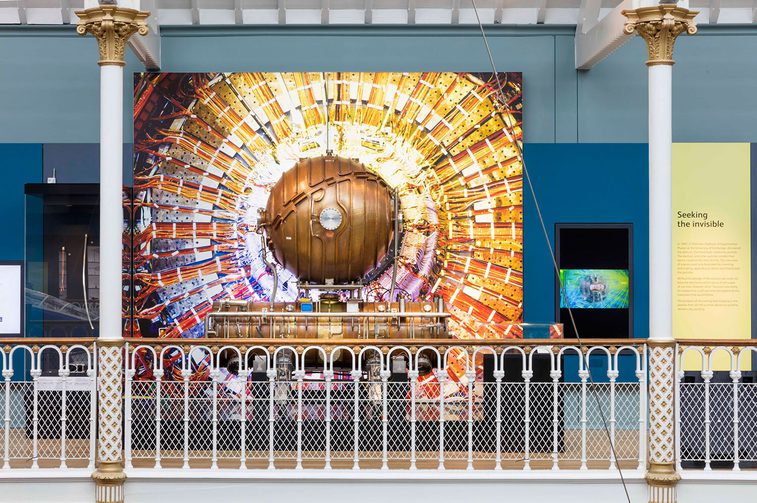 A colour photograph of a museum display about the accelerating cavity. There is a large colour image behind a white metal railing. In between the railing and the image is an example of the copper cavity cylinder. Either side of the display are 2 thin white pillars.