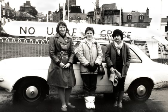 Kristin Barrett and two other women standing in front of a white car, with a cardboard cruise missile with slogan in the background.
