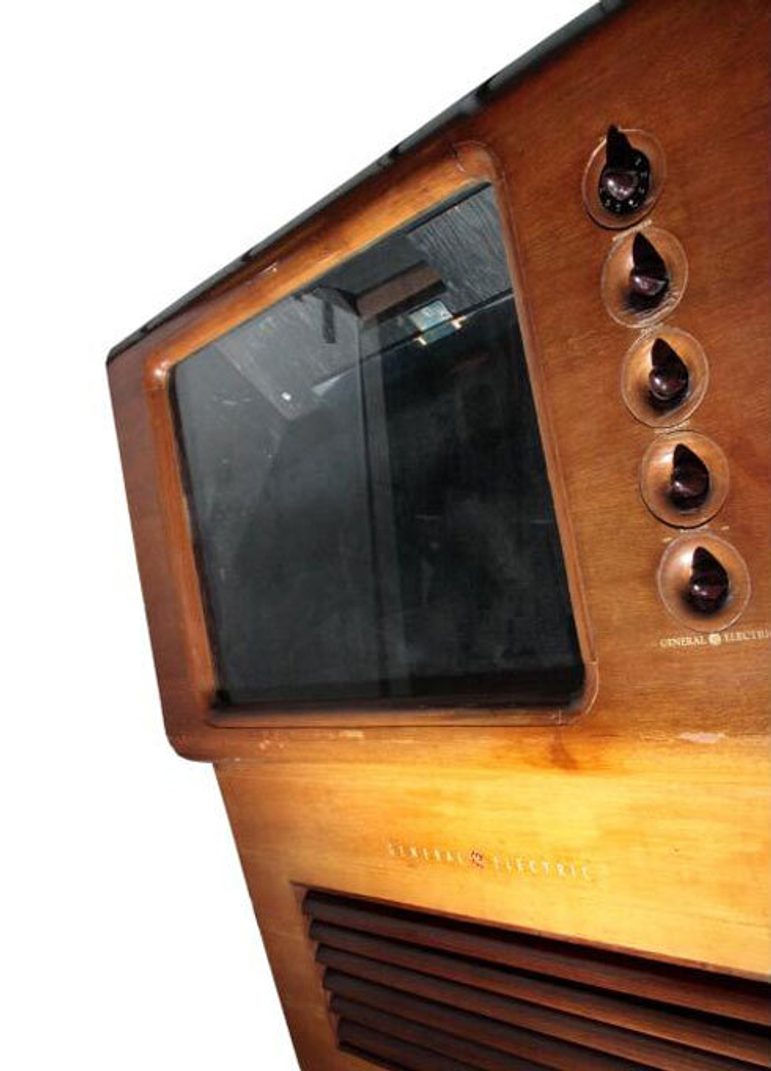 A colour photograph crop of a curved wooden-cased television against a white background. The television screen is to the ;eft and a set of five dials are embedded in the wood to the right. There is a slatted section at the base.