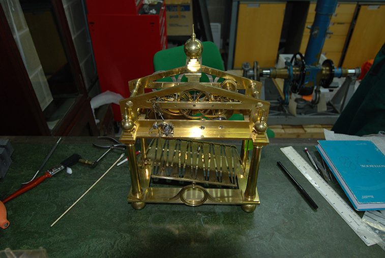 A colour photograph of a brass clock mechanism on top of a workshop bench. There are rulers, pencils and notebooks and other tools also on the bench.