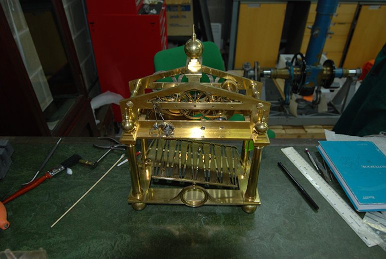 A colour photograph of a brass clock mechanism on top of a workshop bench. There are rulers, pencils and notebooks and other tools also on the bench.