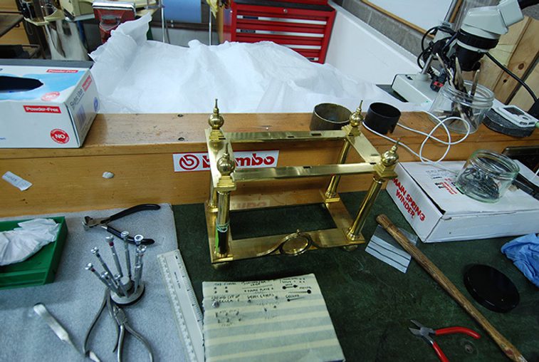 A colour photograph showing a brass frame on a workshop bench. The bench also has tools, screwdrivers, and notes. at the centre is a horizontal wooden bench divider. To the back of the photo is a red tool cabinet and some white material.
