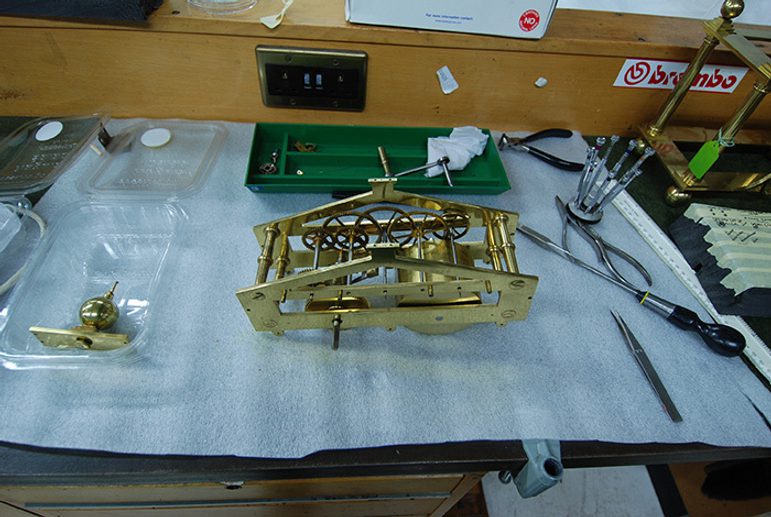 A colour photograph of a brass component on a workbench. The workbench is covered in a protective white cloth, There a specialist screwdrivers and other tools on the bench. There is a wooden bench divider at the back of the image with a set off electrical sockets inset. other brass components are in the periphery of the image.