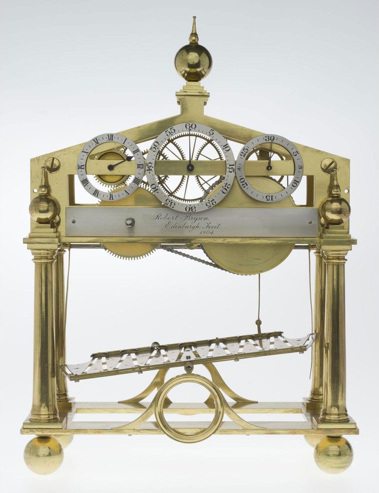 A colour photograph of a brass clock against a white background, with an arch shaped frame and with a central titling horizontal plate. At the top of the clock is an arch mechanical section with several large cogs and wheels. At the very top is a pointed spire. There are 4 columns with brass spheres at the bottom of each.
