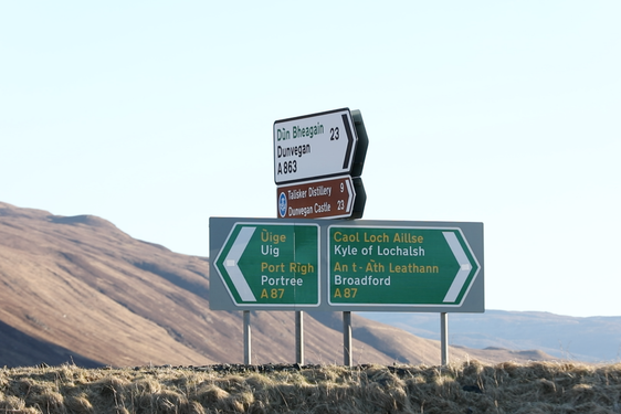 A photograph of four road signs set against a hilly landscape. Two green arrow signs pointing to opposite directions and two signs, one white, the other brown, above the other pointing to the right hand side.
