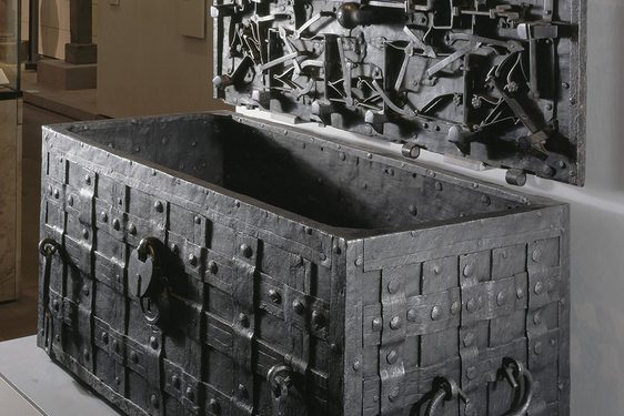 A metal chest decorated with a lattice weave. An open lid is hung on the wall next to the chest. The inside of the lid has multiple metal objects with floral motifs attached to it.