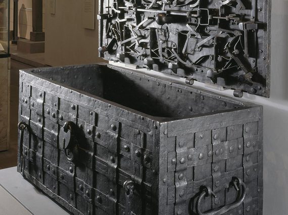 A metal chest decorated with a lattice weave. An open lid is hung on the wall next to the chest. The inside of the lid has multiple metal objects with floral motifs attached to it.
