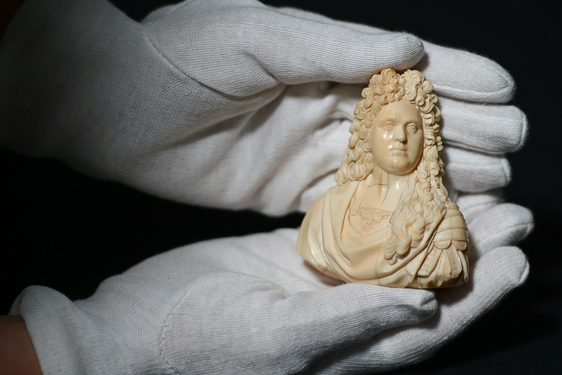 A pair of hands wearing white gloves holding a small ivory statue of a man from the shoulders up with long curly hair and a draped shirt