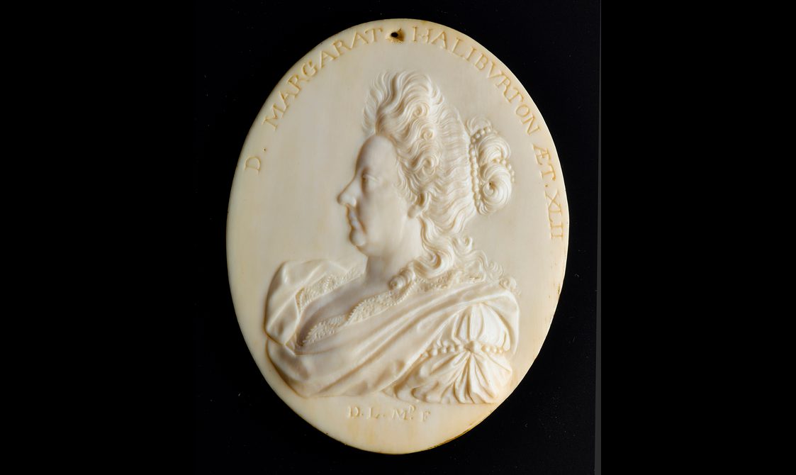 An oval ivory object with a side profile of a woman carved in relief. The woman has her hair pined up and is wearing what appears to be an elaborate dress and there is text around the top of the oval