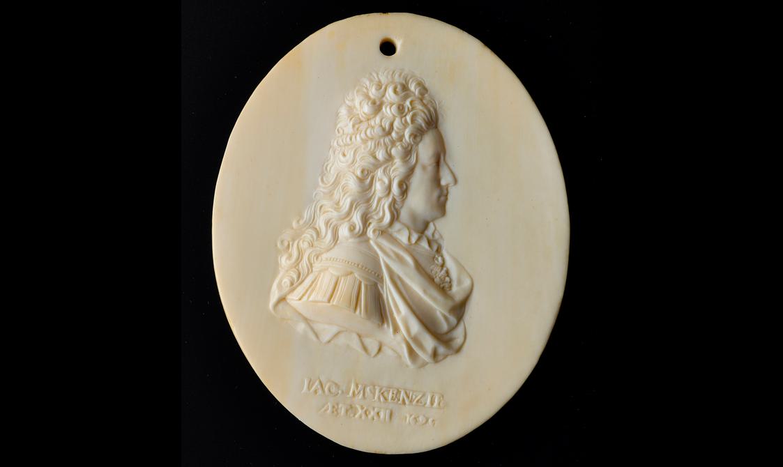 An oval ivory object with a side profile of a man carved in relief. The man has long curly hair and is wearing an elaborate draped shirt and there is text underneath the figure