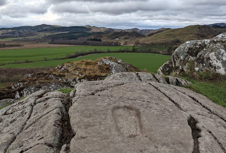 A rock with a footprint imprinted on its surface overlooking green fields and green and brown hills