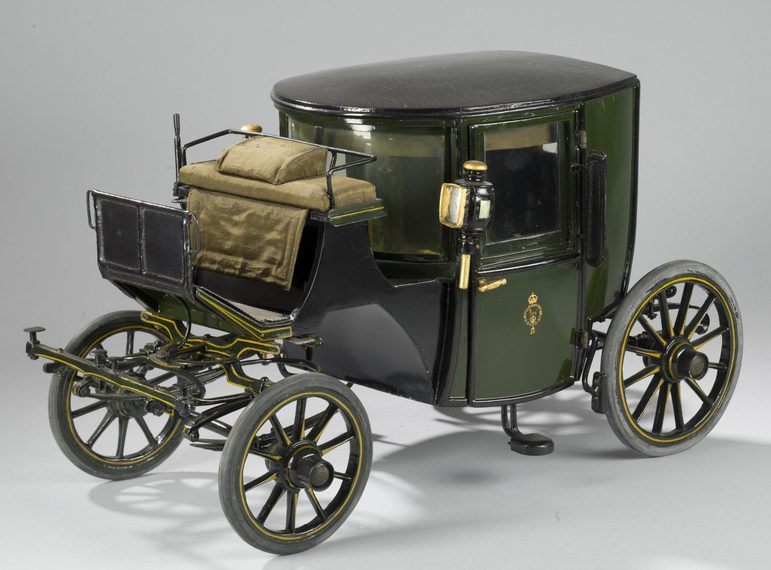 A colour photograph of a model of a victorian horse-drawn carriage with four wheels with pneumatic tyres. The driver section is high-up and outside and the passenger section is a wooden container with two doors.