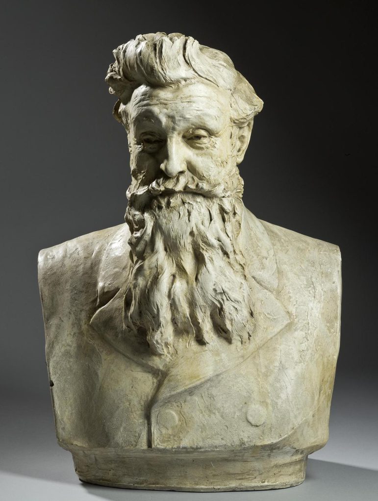 A colour photograph of a marble bust sculpture against a black background. The bust is of a man with swept-back hair, and a long beard. wearing a Victorian double-breasted coat.