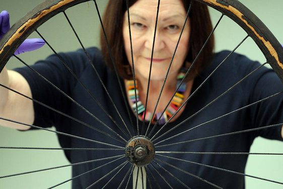 A colour photograph of a woman with dark hair wearing a black top and purple gloves against a gradient green background. She's holding a large wheel in front of her with 30 spokes. It is quite weathered. The rim is surrounded by a degraded rubber.