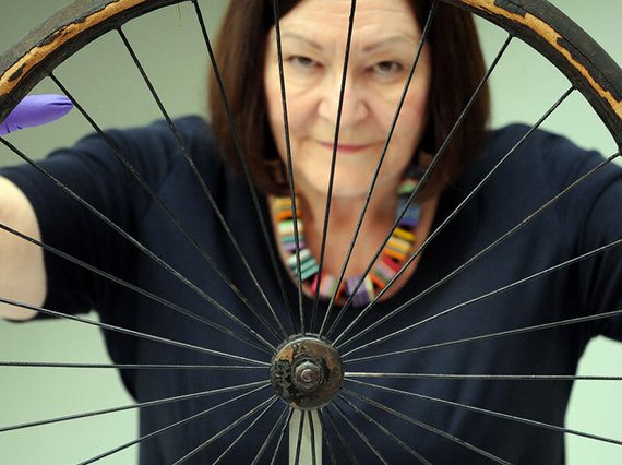 A colour photograph of a woman with dark hair wearing a black top and purple gloves against a gradient green background. She's holding a large wheel in front of her with 30 spokes. It is quite weathered. The rim is surrounded by a degraded rubber.